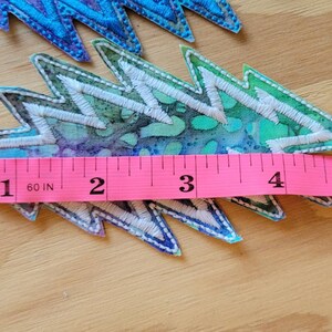 Puede incluir: Un parche de tela de estilo tie-dye colorido con forma de rayo. El parche mide aproximadamente 4 pulgadas de largo y se mide con una cinta m&eacute;trica rosa.