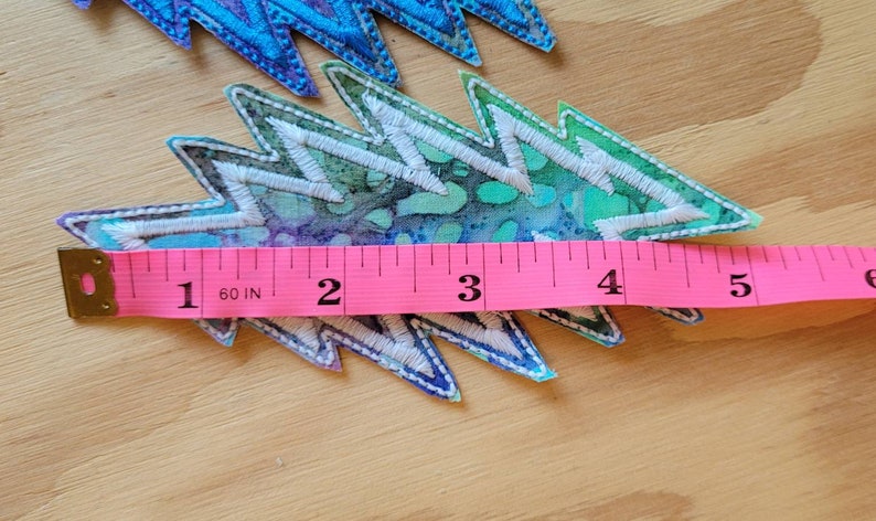 Puede incluir: Un parche de tela de estilo tie-dye colorido con forma de rayo. El parche mide aproximadamente 4 pulgadas de largo y se mide con una cinta m&eacute;trica rosa.