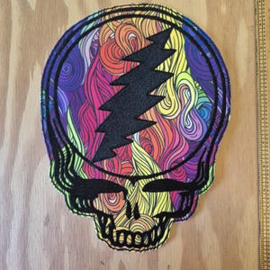 Könnte beinhalten: Ein farbenfroher Aufnäher mit einem Totenkopf-Design und einem Blitz. Der Totenkopf ist schwarz umrandet, mit einem lebendigen Regenbogenwirbelmuster im Inneren. Der Blitz ist schwarz. Der Aufnäher ist etwa 20 cm hoch.