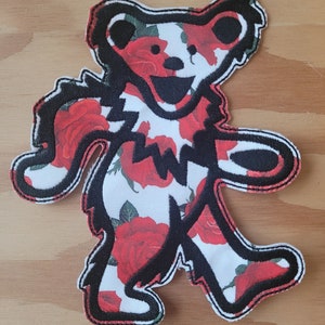 May include: Black and white embroidered patch of a bear with a red rose pattern. The bear is standing with its arms outstretched.