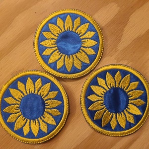 May include: Three embroidered iron-on patches with a sunflower design. The sunflowers are yellow with blue centers and are surrounded by a blue circle with a yellow outline.