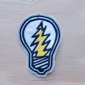 Könnte beinhalten: Ein weißer gestickter Patch in Form einer Glühbirne mit einem gelben Blitz im Inneren. Der Umriss der Glühbirne ist schwarz.