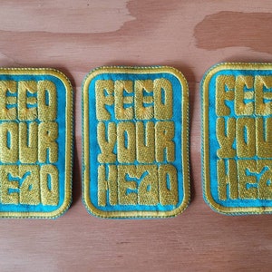 Può includere: Tre toppe ricamate di colore turchese con cuciture gialle. Ogni toppa riporta la scritta "Feed Your Head".