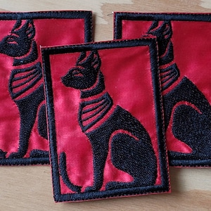 Puede incluir: Tres parches de tela roja con imágenes bordadas en negro de un gato sentado con collar. El gato está estilizado para parecerse a una deidad egipcia.