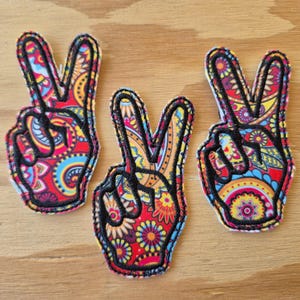 Peut inclure: Trois patchs en forme de signe de paix avec un motif paisley dans des tons de rouge, bleu, jaune et orange. Les patchs sont soulignés de noir.