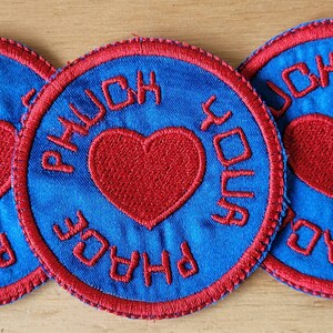 Peut inclure: Un patch brodé bleu et rouge avec un cœur et le texte "PHUCK YOU PHASE".