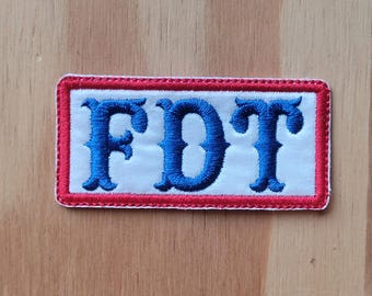 Può includere: Una toppa bianca con un bordo rosso e lettere ricamate blu che compongono la parola "FDT".