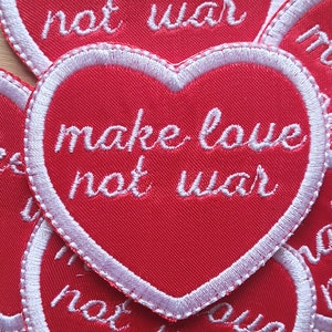 May include: Red heart-shaped patch with white stitching around the edges. The patch says "make love not war" in white embroidery.