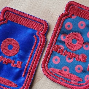 May include: Two embroidered iron-on patches in the shape of a jar. One patch is blue with a red donut and the word "SAMPLE" in red. The other patch is dark blue with a red donut pattern and the word "SAMPLE" in red.