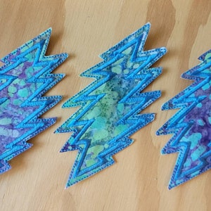 Puede incluir: Tres parches de rayo azul y morado con estampado tie-dye y costuras blancas.