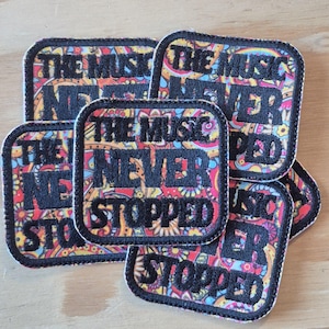 Könnte beinhalten: Mehrere quadratische Aufnäher mit dem Text "THE MUSIC NEVER STOPPED" in Schwarz, weiß umrandet. Die Aufnäher haben ein farbenfrohes Paisley-Design in Rot, Gelb, Blau und Orange. Die Aufnäher liegen auf einer hellbraunen Oberfläche.