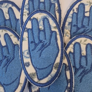 Puede incluir: Una colección de parches bordados de forma ovalada con un gesto de mano azul sobre un fondo blanco con un patrón floral azul. Cada parche tiene un borde cosido en azul y muestra una mano con el dedo índice y el meñique extendidos.