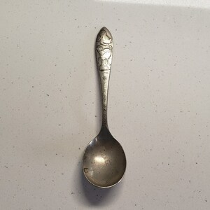 May include: A silver spoon with a cartoon character design on the handle. The character is wearing a hat and holding a bucket.