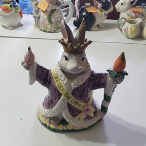May include: A white rabbit teapot with a purple robe and a crown. The teapot is decorated with flowers and has a carrot-shaped spout. The teapot is labeled "Peter the Great".