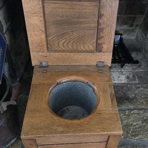 May include: An antique wooden commode with a hinged lid. The commode has a circular opening with a textured metal interior. The wood has a natural finish, and the piece is set on short legs. The lid is open, revealing the interior.