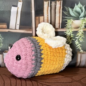 May include: A plush crochet bee toy featuring a pink head, a gray midsection, and a yellow body with white wings. The toy has two small black plastic eyes on the sides of the pink head and a white tail end, crafted from soft, textured yarn.