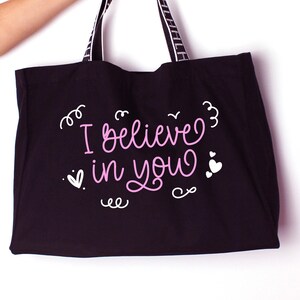 K&ouml;nnte beinhalten: Schwarze Tragetasche mit wei&szlig;en Henkeln und dem Text "I believe in you" in rosa Schrift mit wei&szlig;en Herzen und Schn&ouml;rkeln.