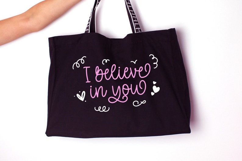 K&ouml;nnte beinhalten: Schwarze Tragetasche mit wei&szlig;en Henkeln und dem Text "I believe in you" in rosa Schrift mit wei&szlig;en Herzen und Schn&ouml;rkeln.