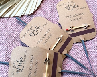 May include: Three brown paper sparklers with a gray stick and a small wooden match attached. The paper has the text "Lasst unsere Liebe leuchten" which translates to "Let our love shine" and the date "01.07.2023" and the names "Tine & Bernd".