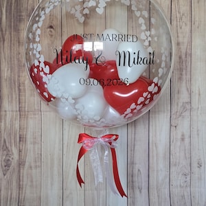 K&ouml;nnte beinhalten: Ein klarer Bubble-Ballon, gef&uuml;llt mit roten und wei&szlig;en herzf&ouml;rmigen Luftballons. Der Ballon ist mit wei&szlig;en Herzmustern und den Worten "JUST MARRIED" sowie Namen und einem Datum verziert. Unten ist ein rot-wei&szlig;es Band gebunden.