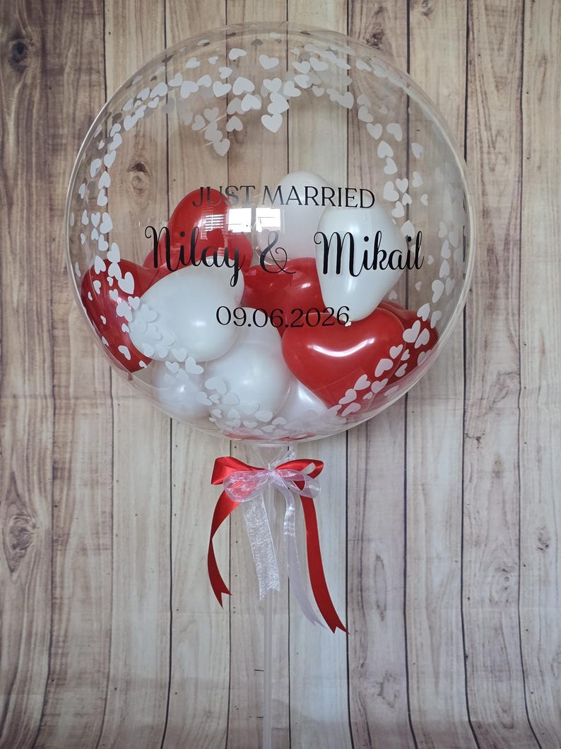 K&ouml;nnte beinhalten: Ein klarer Bubble-Ballon, gef&uuml;llt mit roten und wei&szlig;en herzf&ouml;rmigen Luftballons. Der Ballon ist mit wei&szlig;en Herzmustern und den Worten "JUST MARRIED" sowie Namen und einem Datum verziert. Unten ist ein rot-wei&szlig;es Band gebunden.