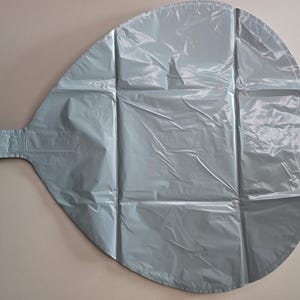 Op de afbeelding: Een grote, lichtblauwe ballon in een druppelvorm, gemaakt van een gekreukeld, glanzend materiaal. De ballon heeft een rechthoekig lipje dat zich vanaf &eacute;&eacute;n uiteinde uitstrekt en is verdeeld in secties door verticale en horizontale naden.