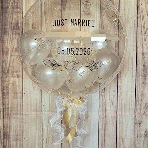 May include: A large clear balloon containing smaller white balloons with gold flakes. The exterior features black text reading JUST MARRIED and 05.05.2026, along with a graphic of two hearts and leafy branches. A white and gold ribbon is tied at the base.