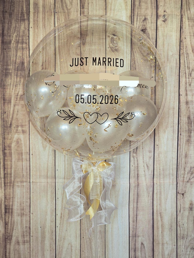 May include: A large clear balloon containing smaller white balloons with gold flakes. The exterior features black text reading JUST MARRIED and 05.05.2026, along with a graphic of two hearts and leafy branches. A white and gold ribbon is tied at the base.