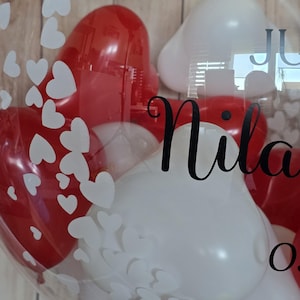K&ouml;nnte beinhalten: Ein klarer Ballon gef&uuml;llt mit roten und wei&szlig;en herzf&ouml;rmigen Ballons. Der Ballon hat wei&szlig;e herzf&ouml;rmige Aufkleber und den Namen "Nila" in schwarzer Schrift. Der Hintergrund ist eine hellbraune Holzoberfl&auml;che.