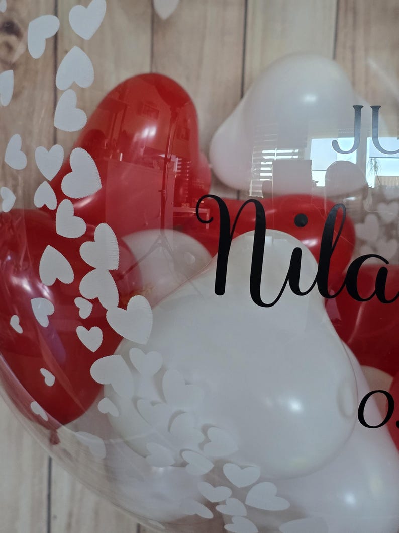 K&ouml;nnte beinhalten: Ein klarer Ballon gef&uuml;llt mit roten und wei&szlig;en herzf&ouml;rmigen Ballons. Der Ballon hat wei&szlig;e herzf&ouml;rmige Aufkleber und den Namen "Nila" in schwarzer Schrift. Der Hintergrund ist eine hellbraune Holzoberfl&auml;che.