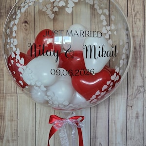K&ouml;nnte beinhalten: Ein durchsichtiger Bubble-Ballon, gef&uuml;llt mit roten und wei&szlig;en Herz- und runden Luftballons. Der Ballon tr&auml;gt die Aufschrift "Just Married", die Namen "Nilay & Mikail" und das Datum "09.06.2026". Unten ist ein rot-wei&szlig;es Band befestigt.