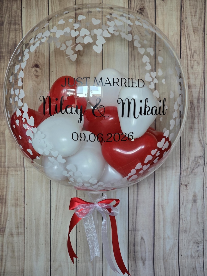 K&ouml;nnte beinhalten: Ein durchsichtiger Bubble-Ballon, gef&uuml;llt mit roten und wei&szlig;en Herz- und runden Luftballons. Der Ballon tr&auml;gt die Aufschrift "Just Married", die Namen "Nilay & Mikail" und das Datum "09.06.2026". Unten ist ein rot-wei&szlig;es Band befestigt.