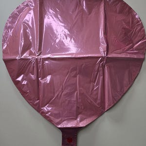 Op de afbeelding: Een roze hartvormige ballon gemaakt van glanzend, gekreukeld materiaal. De ballon heeft een ventiel aan de onderkant om op te blazen en een kleine rode cirkel bij het ventiel. De ballon is ontworpen voor feesten en evenementen.
