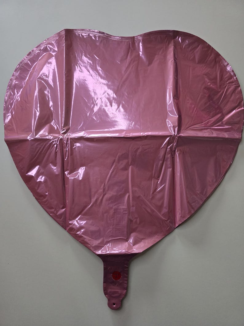 Op de afbeelding: Een roze hartvormige ballon gemaakt van glanzend, gekreukeld materiaal. De ballon heeft een ventiel aan de onderkant om op te blazen en een kleine rode cirkel bij het ventiel. De ballon is ontworpen voor feesten en evenementen.