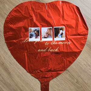 Op de afbeelding: Een grote, rode hartvormige ballon met drie kleine vierkante foto's eraan bevestigd. De ballon heeft de tekst "Love you to the moon and back" in cursief schrift. De ballon is gemaakt van een glanzend, reflecterend materiaal.