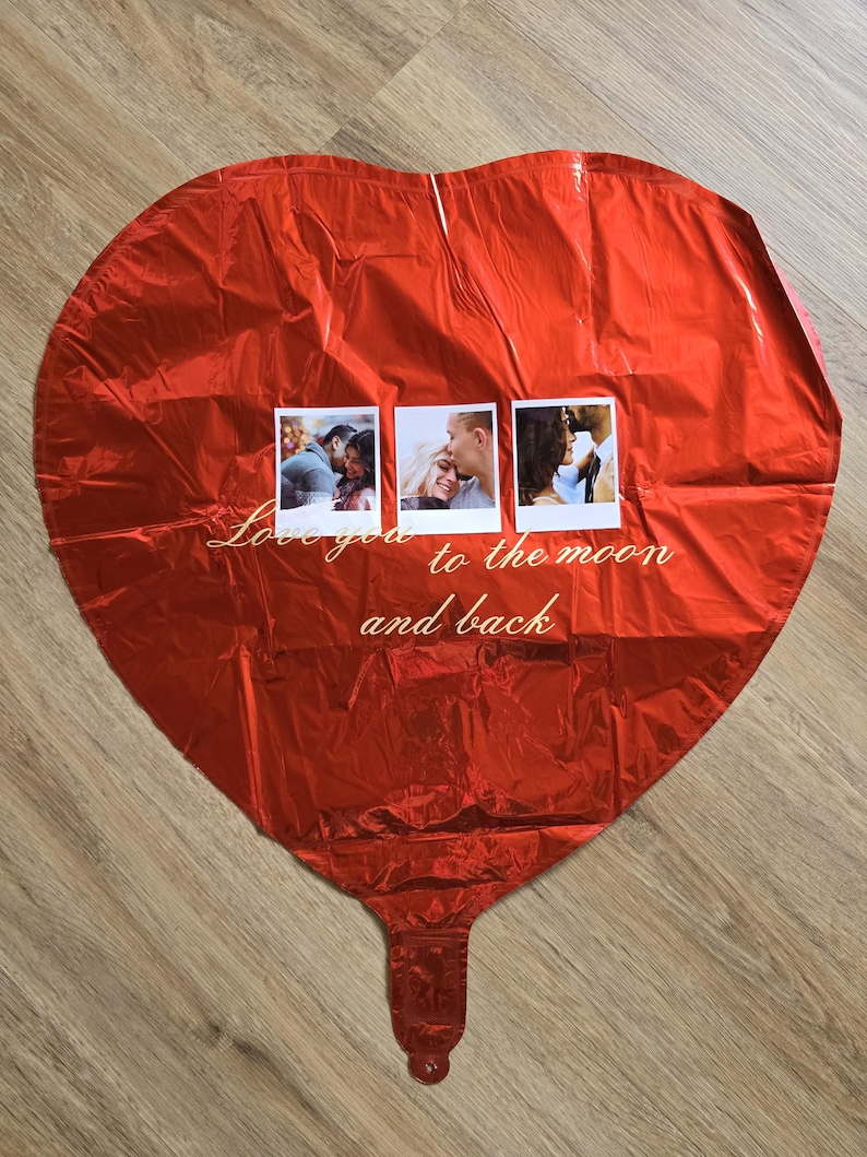 Op de afbeelding: Een grote, rode hartvormige ballon met drie kleine vierkante foto's eraan bevestigd. De ballon heeft de tekst "Love you to the moon and back" in cursief schrift. De ballon is gemaakt van een glanzend, reflecterend materiaal.