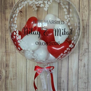 K&ouml;nnte beinhalten: Ein klarer Bubble-Ballon, gef&uuml;llt mit roten und wei&szlig;en herzf&ouml;rmigen Luftballons. Der Ballon ist mit wei&szlig;en Herzmustern und dem Text "Just Married" mit Namen und Datum verziert. Unten ist ein rot-wei&szlig;es Band gebunden.