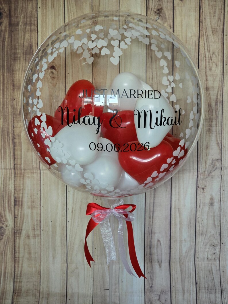 K&ouml;nnte beinhalten: Ein klarer Bubble-Ballon, gef&uuml;llt mit roten und wei&szlig;en herzf&ouml;rmigen Luftballons. Der Ballon ist mit wei&szlig;en Herzmustern und dem Text "Just Married" mit Namen und Datum verziert. Unten ist ein rot-wei&szlig;es Band gebunden.