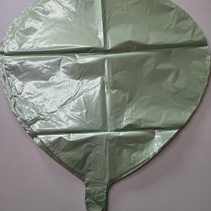Op de afbeelding: Een grote, hartvormige ballon in een gedempte groene kleur. De ballon is gemaakt van een glanzend, gekreukt materiaal en heeft een gevouwen basis met een kleine, knoopachtige sluiting. De ballon is ontworpen voor feesten en evenementen.