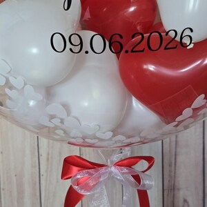 K&ouml;nnte beinhalten: Ein klarer Ballon gef&uuml;llt mit wei&szlig;en und roten herzf&ouml;rmigen Luftballons. Der Ballon ist mit wei&szlig;en Herzmustern und dem Datum 09.06.2026 verziert. Am Boden des Ballons ist ein rot-wei&szlig;es Band gebunden.