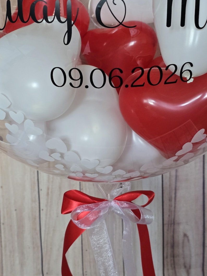 K&ouml;nnte beinhalten: Ein klarer Ballon gef&uuml;llt mit wei&szlig;en und roten herzf&ouml;rmigen Luftballons. Der Ballon ist mit wei&szlig;en Herzmustern und dem Datum 09.06.2026 verziert. Am Boden des Ballons ist ein rot-wei&szlig;es Band gebunden.