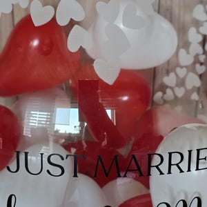 K&ouml;nnte beinhalten: Eine transparente Box gef&uuml;llt mit roten und wei&szlig;en herzf&ouml;rmigen Luftballons. Die Worte "JUST MARRIED" sind in schwarzer Schrift auf die Box gedruckt. Wei&szlig;e herzf&ouml;rmige Ausschnitte sind auf der Box angebracht.