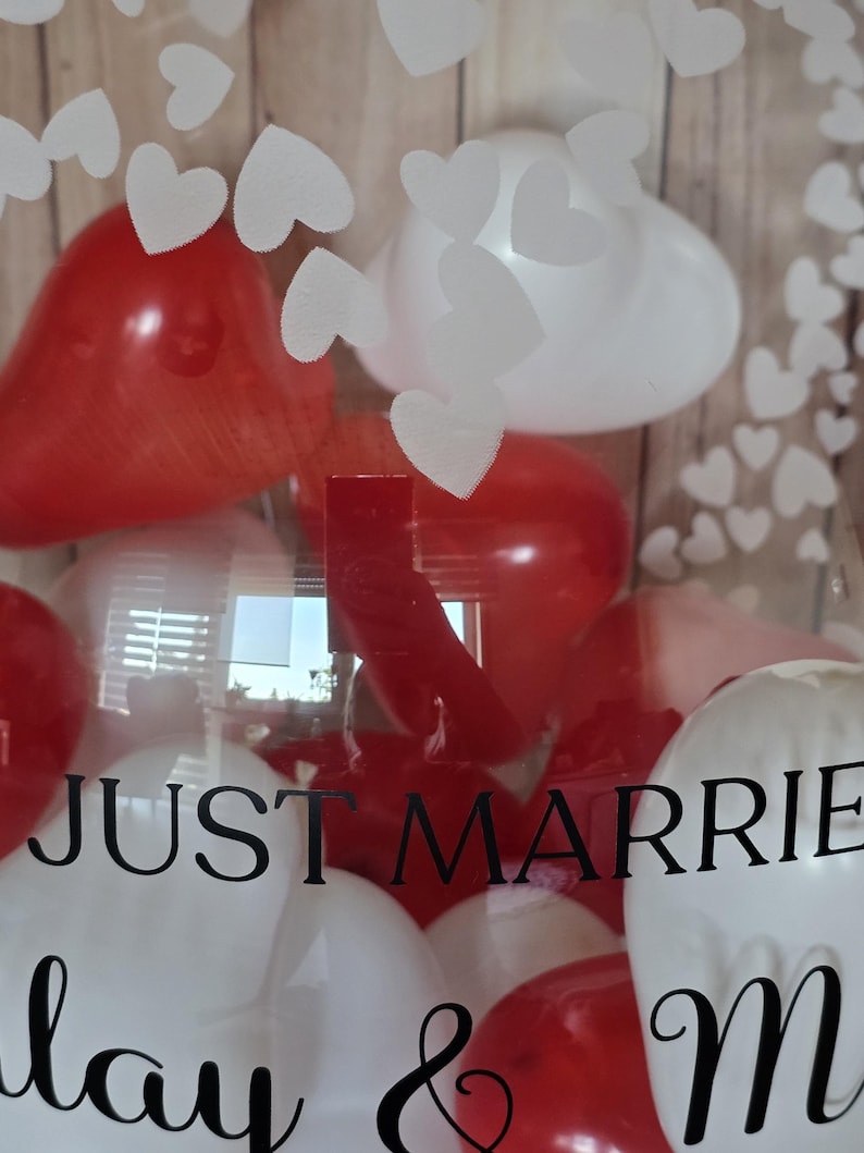 K&ouml;nnte beinhalten: Eine transparente Box gef&uuml;llt mit roten und wei&szlig;en herzf&ouml;rmigen Luftballons. Die Worte "JUST MARRIED" sind in schwarzer Schrift auf die Box gedruckt. Wei&szlig;e herzf&ouml;rmige Ausschnitte sind auf der Box angebracht.