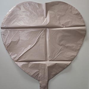 Op de afbeelding: Een grote, hartvormige ballon in een gedempte, stoffige roze kleur. De ballon heeft een glanzende, metallic afwerking en is gekreukt met vouwen. De ballon heeft een mondstuk aan de onderkant om op te blazen.