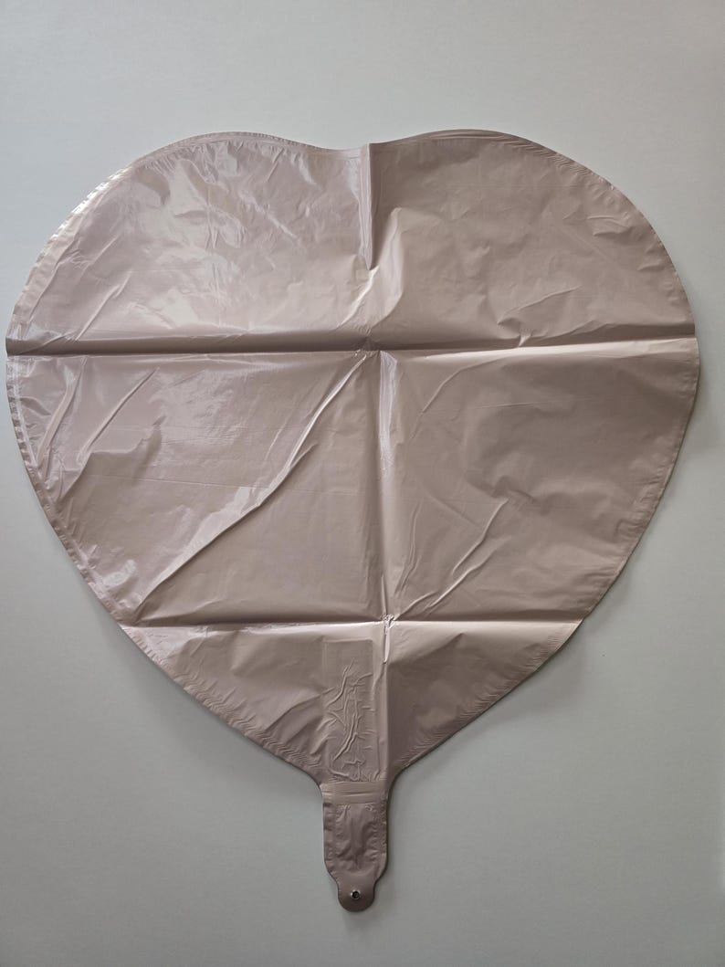 Op de afbeelding: Een grote, hartvormige ballon in een gedempte, stoffige roze kleur. De ballon heeft een glanzende, metallic afwerking en is gekreukt met vouwen. De ballon heeft een mondstuk aan de onderkant om op te blazen.
