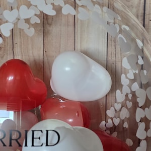 K&ouml;nnte beinhalten: Ein klarer Ballon gef&uuml;llt mit roten und wei&szlig;en herzf&ouml;rmigen Luftballons. Der Ballon hat ein Muster aus wei&szlig;en Herzen auf der Oberfl&auml;che. Das Wort "Married" ist teilweise auf dem Ballon zu sehen.