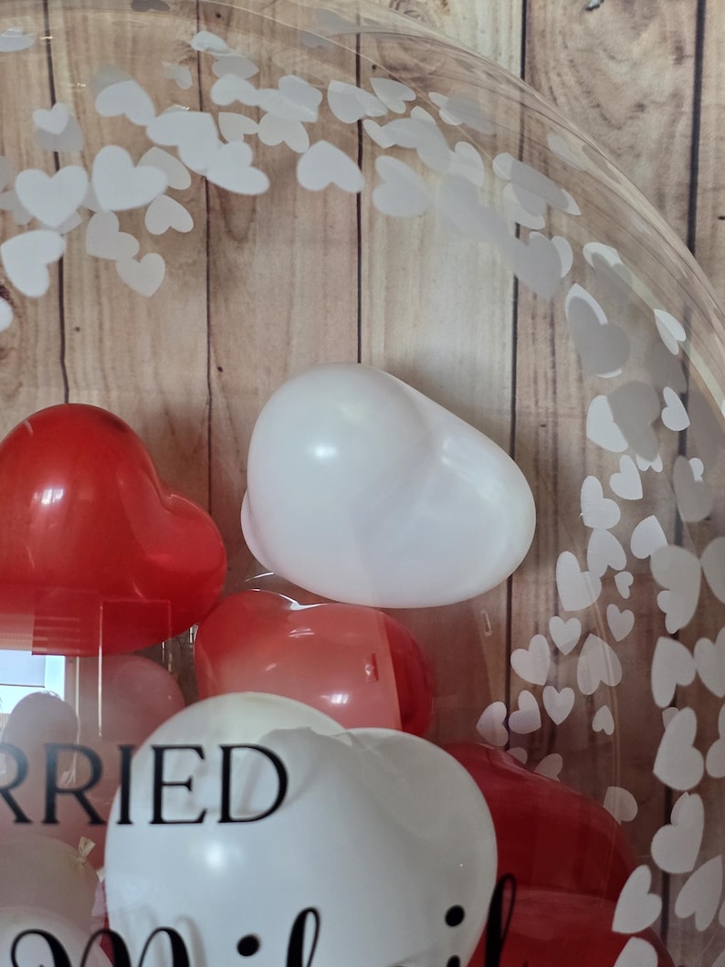 K&ouml;nnte beinhalten: Ein klarer Ballon gef&uuml;llt mit roten und wei&szlig;en herzf&ouml;rmigen Luftballons. Der Ballon hat ein Muster aus wei&szlig;en Herzen auf der Oberfl&auml;che. Das Wort "Married" ist teilweise auf dem Ballon zu sehen.