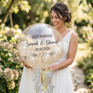 May include: A large, clear, spherical balloon filled with smaller white balloons and gold confetti. The exterior features black text reading JUST MARRIED, Sarah & David, 05.05.2026, and a decorative heart and leaf graphic. A silver and gold ribbon is tied at the base.