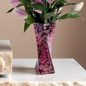 Könnte beinhalten: Ein lebendiges Blumenarrangement mit rosa Lilien in einer dekorativen Vase. Die Vase hat ein einzigartiges, eckiges Design mit einem tiefroten und goldenen Muster. Die Lilien blühen voll, mit grünen Blättern, die einen Kontrast bilden. Die Vase ist etwa 25 cm hoch.