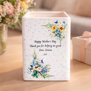 May include: A white square vase with a floral design and the text "Happy Mother's Day." The vase is decorated with blue, yellow, and orange flowers and butterflies. A bouquet of flowers and a gift box are also on the table.